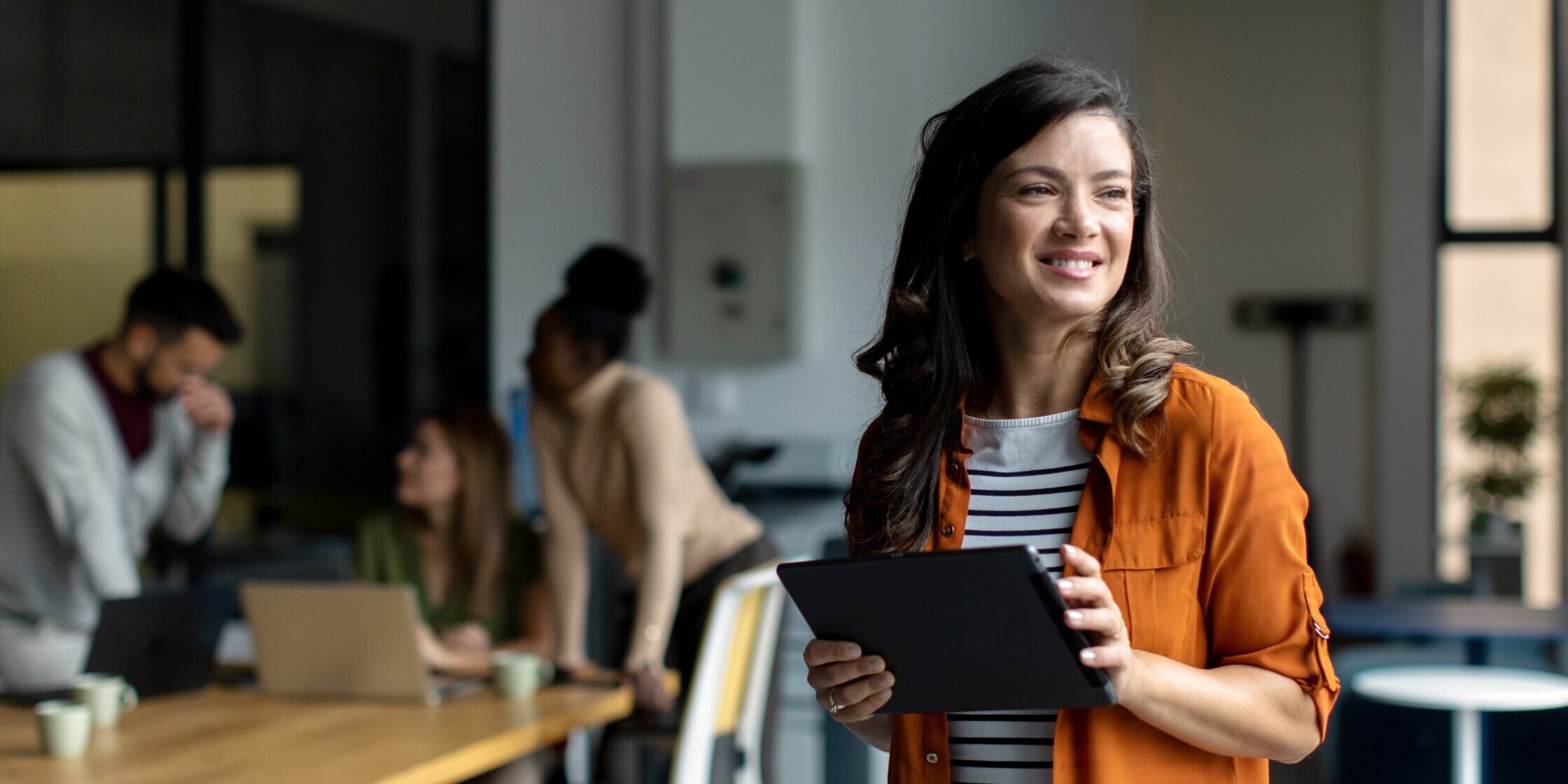 Mid adult female businesswoman standing and walking through modern office while her colleagues working and brainstorming